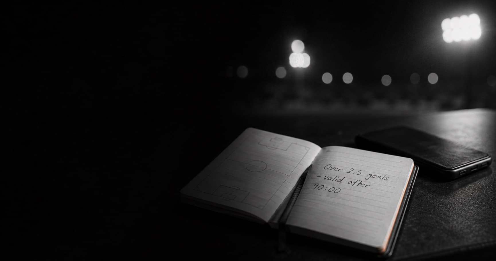 Notebook open on a pitch diagram with a handwritten market rule, phone face-down, night stadium lights